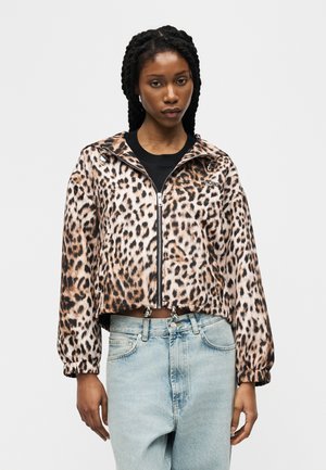 Kobieta z warkoczami, ubrana w kurtkę na zamek z nadrukiem w panterkę oraz jasnoniebieskie dżinsy z wysokim stanem na jednolitym tle.