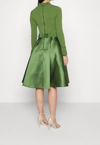 Vestido verde con un top ajustado de manga larga y una falda satinada acampanada. Cuenta con un cierre de cremallera y un lazo decorativo en la cintura.