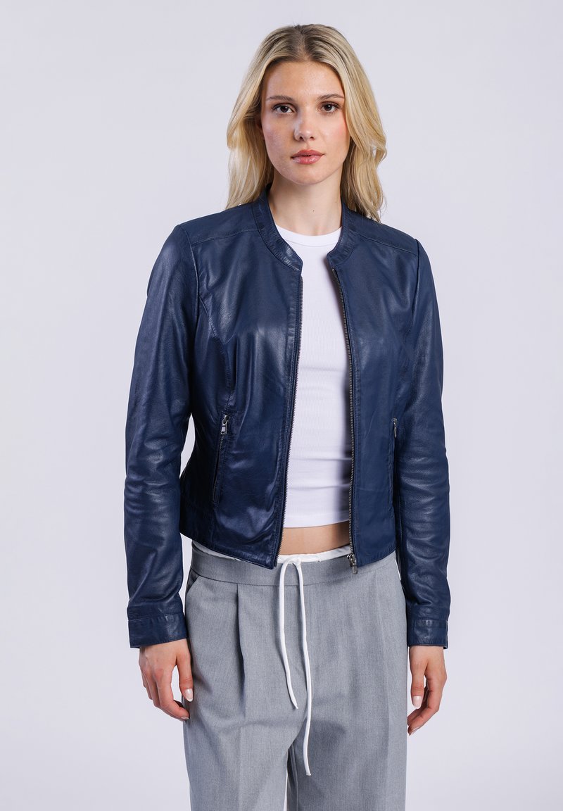 Veste en cuir bleu marine au design ajusté, col rond et poches zippées ; portée sur un haut blanc et associée à un pantalon gris.