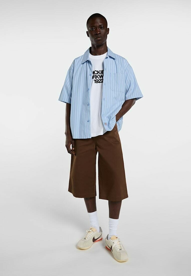 Mann steht in braunen knielangen Shorts, weißem T-Shirt mit Grafik, hellblauem kurzärmeligem Hemd mit Knöpfen, weißen Socken und beigen Sneakern.