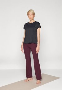 Svart atletisk topp med korta ärmar och böjda sömmar, kombinerad med vinröda utsvängda byxor. Slät tyg, avslappnad passform, stående på en beige matta.