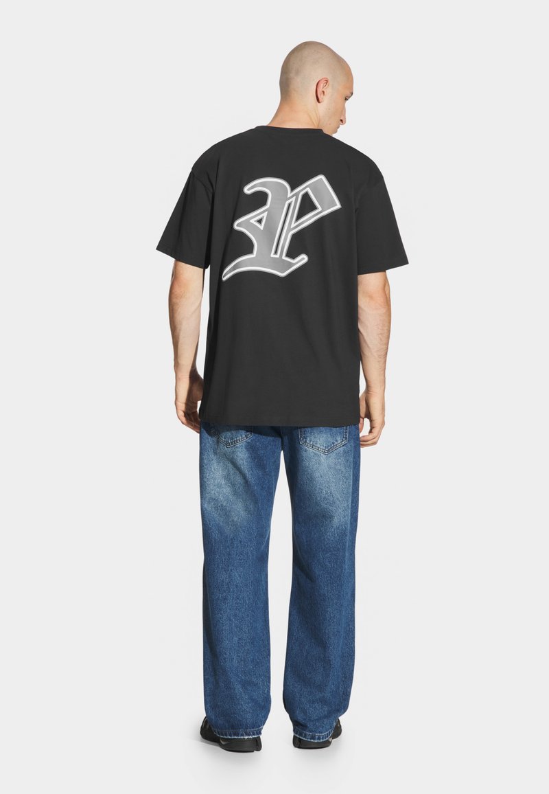 Czarna bawełniana koszulka w oversize'owym kroju z krótkim rękawem. Na plecach znajduje się duży biały graficzny logotyp. Zestawiona z jasnymi niebieskimi dżinsami.