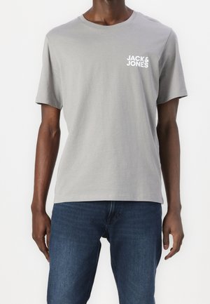 Mężczyzna ubrany w jasnoszary t-shirt Jack & Jones i niebieskie dżinsy stoi na białym tle.