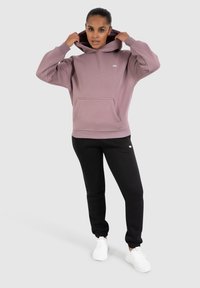 Femme ajustant la capuche d'un hoodie mauve, portant un pantalon de jogging noir et des baskets blanches, se tenant devant un fond neutre.