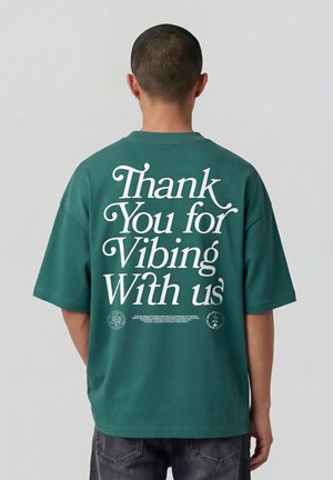 Persoon met kort haar die een groene oversized T-shirt draagt met witte tekst "Thank You for Vibing With us" op de achterkant.