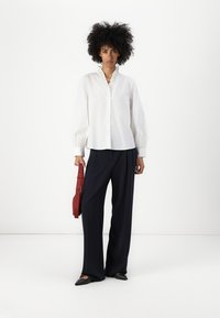 Camisa blanca de botones con cuello rizado, mangas amplias, combinada con pantalones de pierna ancha en navy y un bolso rojo de tirante. Zapatos planos negros.