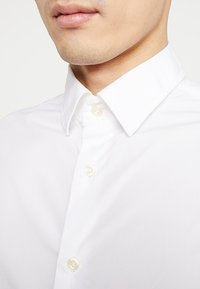 Biała koszula z kołnierzykiem typu button-down, wykonana z gładkiej bawełnianej tkaniny. Posiada kremowe guziki i zbliżenie na dekolt.