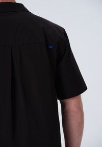 Schwarzes Kurzarmhemd mit Kragen, Falten-Detail am Rücken und einem kleinen blauen Akzent nahe der Schulter. Glatte Textur.