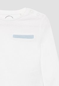 Witte babyshirt met lange mouwen, met een blauwe borstzak en twee drukknoopjes op de linkerschouder voor eenvoudige aankleding.