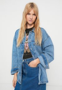 Denim jacket in light blue with a relaxed fit, featuring two chest pockets, contrast stitching, and folded sleeves, worn over a graphic t-shirt.