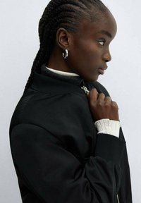 Schwarze Bomberjacke mit gerippten Bündchen, hohem Kragen und silbernem Reißverschluss. Über einem weißen, strukturierten Pullover getragen. Das Model hat geflochtenes Haar.