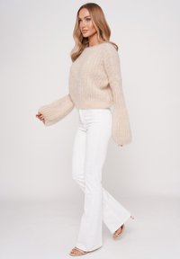 Beige knit sweater with ribbed texture, wide sleeves, paired with white flared jeans and nude sandals, set against a plain background.