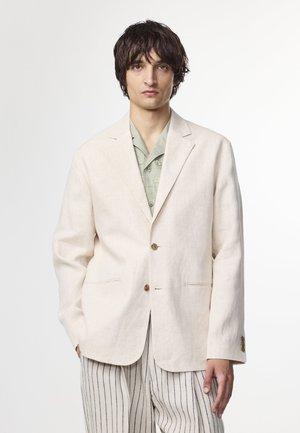 Beige linneblazer med en knappstängning, spetsiga slag och två framfickor. Matchad med en ljusgrön mönstrad skjorta och randiga byxor.