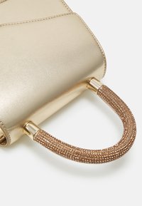 Gros plan d'un sac à main doré avec une anse courbe ornée de petits cristaux dorés et un corps en cuir lisse.