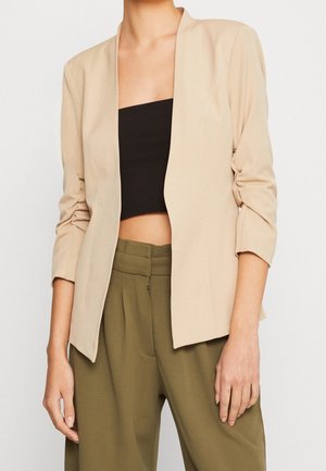 Kvinde iført en beige blazer over en sort tube top og olivengrønne højtaljede bukser, stående med afslappede arme.