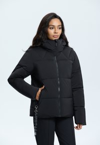 Schwarze Puffjacke mit Kapuze, ausgestattet mit einem Reißverschluss vorne, geometrischer Steppung und Seitentaschen. Mit einem dekorativen Hardware-Detail akzentuiert.
