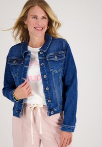 Vrouw die glimlacht, gekleed in een blauwe spijkerjasje over een wit shirt met roze tekst, lichtroze broeken met een strikkoord, gouden hoop earrings.