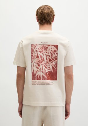 Blonde Person trägt ein weißes T-Shirt mit rotem Bambusblattdruck und kleinem Text auf dem Rücken, steht vor einem einfarbigen weißen Hintergrund.