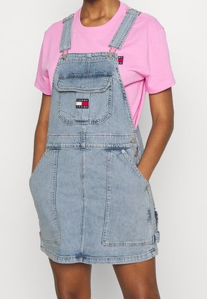 Person trägt ein hellblaues Jeans-Latzkleid mit Vordertasche und ein rosa Kurzarmshirt, Hände in den Seitentaschen, sichtbare Tommy Jeans-Logos.