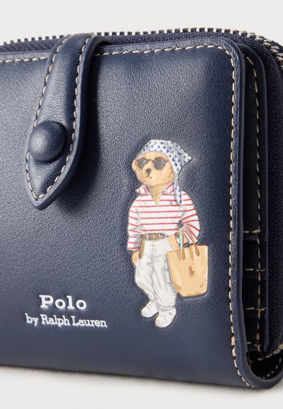 Πορτοφόλι από δέρμα σε ναυτικό μπλε με ραμμένες άκρες, κούμπωμα με κουμπί και απεικόνιση αρκούδας που φοράει γυαλιά ηλίου, ριγέ πουκάμισο και κρατάει μπεζ τσάντα.