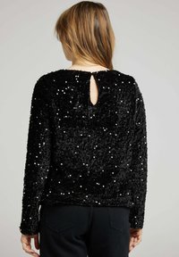 Top en sequins noirs avec une texture douce, présentant des manches longues et un détail en forme de trou de serrure au dos. Des sequins scintillants créent un motif à pois.