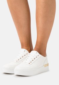 Des baskets blanches avec un dessus texturé, un bout rond et des œillets dorés. Elles possèdent une semelle blanche épaisse et un subtil accent doré au talon.