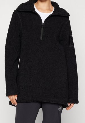 Femme portant un pull polaire noir texturé avec une fermeture éclair au quart, par-dessus une chemise blanche et un pantalon noir avec un petit logo près du genou.
