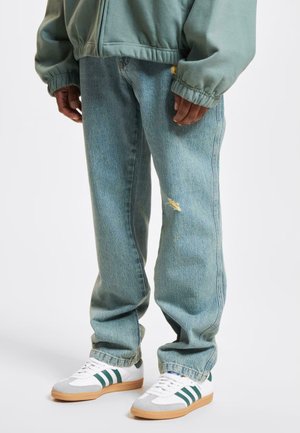 Világoskék, laza szabású farmernadrág, rojtos részletekkel, fehér sportcipőkkel párosítva, zöld csíkokkal és szürke velúr orral.