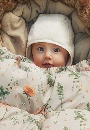 Baby with wide eyes wearing a white hat, wrapped in a floral patterned blanket and surrounded by soft beige fabric.