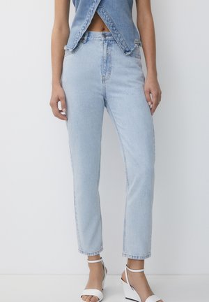 Femme portant un jean taille haute bleu clair, un haut en denim bleu court et des sandales compensées blanches, debout devant un fond blanc uni.