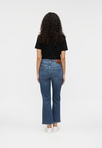 Σκούρα μπλε τζιν με καμπάνα, ψηλόμεσα, με πίσω τσέπες και ταμπά δερμάτινο λογότυπο· συνδυασμένα με μαύρο κοντομάνικο τοπ.