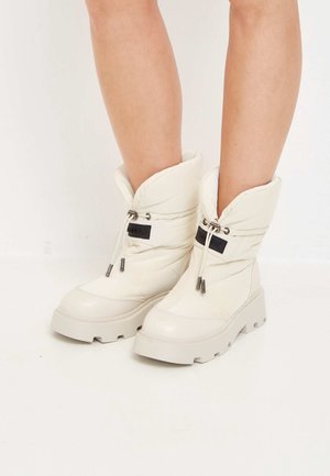 Jambes portant des bottes d'hiver écrues mi-mollet avec des semelles épaisses et des cordons ajustables sur fond blanc.