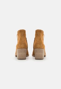 Pár hnědých semišových kotníkových bot s blokovým podpatkem, vzhledem zezadu na bílém pozadí.