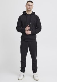 Sudadera negra con capucha y cordones, combinada con pantalones jogger a juego. Ambos tienen una textura suave; el modelo lleva zapatillas blancas.