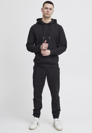 Black hooded sweatshirt with drawstrings, paired with matching joggers. Both have a smooth texture; model wears white sneakers.