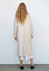 Mujer con cabello largo y ondulado de color rubio, vistiendo un abrigo largo beige sobre una falda blanca y zapatos negros, de pie contra una pared blanca lisa.