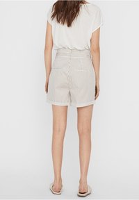 Žena oblečená v bílých krátkých kalhotách s tenkými vertikálními pruhy, zasunutém bílém tričku s krátkým rukávem a béžových pantoflích, stojí čelem otočena pryč.