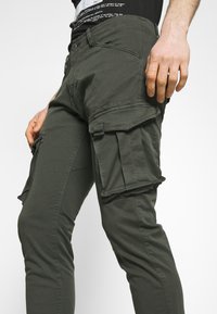 Oliwkowe spodnie cargo o dopasowanym kroju, z wieloma kieszeniami po bokach i gładką teksturą materiału. Zapinane na guziki i suwak z przodu.