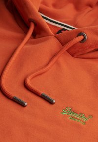 Orange hoodie featuring a drawstring with metal tips, a logo in green embroidery, and a soft texture. Showcase of casual apparel design.