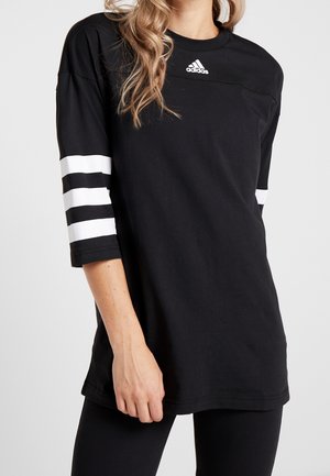 Kvinde iført en sort Adidas-shirt med albuelange ærmer og hvide tre striber på armene, stående mod en ensfarvet baggrund.