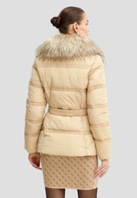 Beige polstret jakke med krave i faux fur, indsnøret talje og vandret sømdetaljering. Sammen med et mønstret nederdel.