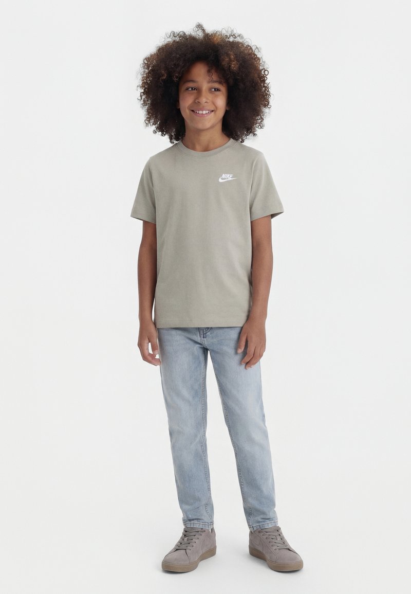 Jeune personne aux cheveux bouclés portant un t-shirt Nike vert clair, un jean bleu clair et des baskets grises, debout et souriant.