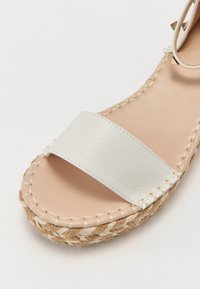 Sandale beige à bout ouvert avec une lanière en cuir blanc et une semelle plateforme en jute tressé, dotée de détails de bride de cheville cloutée.