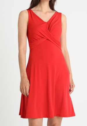 Kvinde iført en ærmeløs rød knælængde kjole med snoet frontdetalje og en V-udskæring på en ensfarvet hvid baggrund.