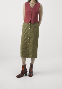 Kobieta w bezrękawnikowej, czerwonej kamizelce na guziki, oliwkowej, midi spódnicy na guziki oraz brązowych botkach za kostkę z rzeźbionymi obcasami.