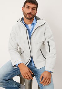 Weiße wasserdichte Jacke mit schwarzen Akzenten, ausgestattet mit einer Kapuze, Reißverschlusstaschen und lockerem Schnitt, kombiniert mit einem hellblauen Shirt und Jeans.