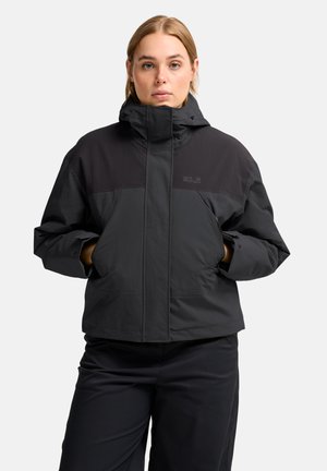 Donna con capelli chiari che indossa una giacca invernale nera con cappuccio e pantaloni neri, mani nelle tasche della giacca, sfondo neutro.