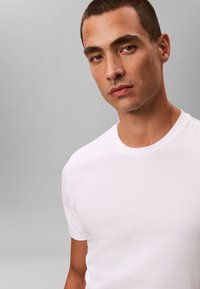Jeune homme aux cheveux courts portant un t-shirt blanc uni à col rond sur un fond gris, regardant légèrement vers la caméra.