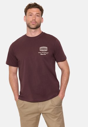Homme aux cheveux courts et bouclés portant un t-shirt marron « Raw & Rugged Adventures » et un pantalon beige, debout les mains dans les poches.