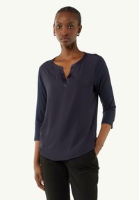 Marineblaue Bluse mit V-Ausschnitt und Dreiviertelärmeln. Glattes Material mit seitlichen Schlitzen für Bewegungsfreiheit. Minimalistisches Design.